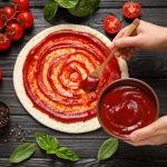 Woman,Spreading,Tomato,Sauce,Onto,Pizza,Crust,On,Dark,Wooden